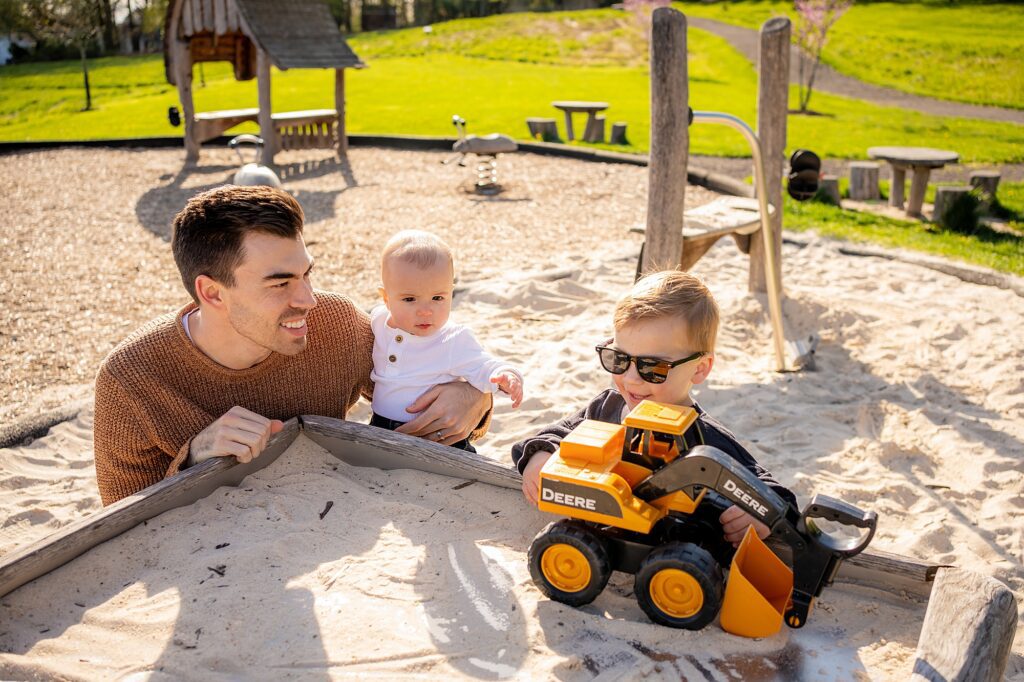 A young man and two small children play with a toy bulldozer in a sandbox at a sunny park.