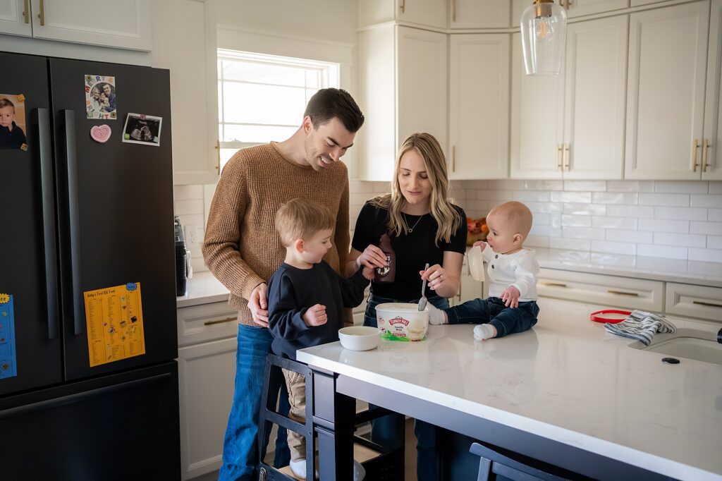 A family in a kitchen, with parents feeding two young children sitting at a counter.