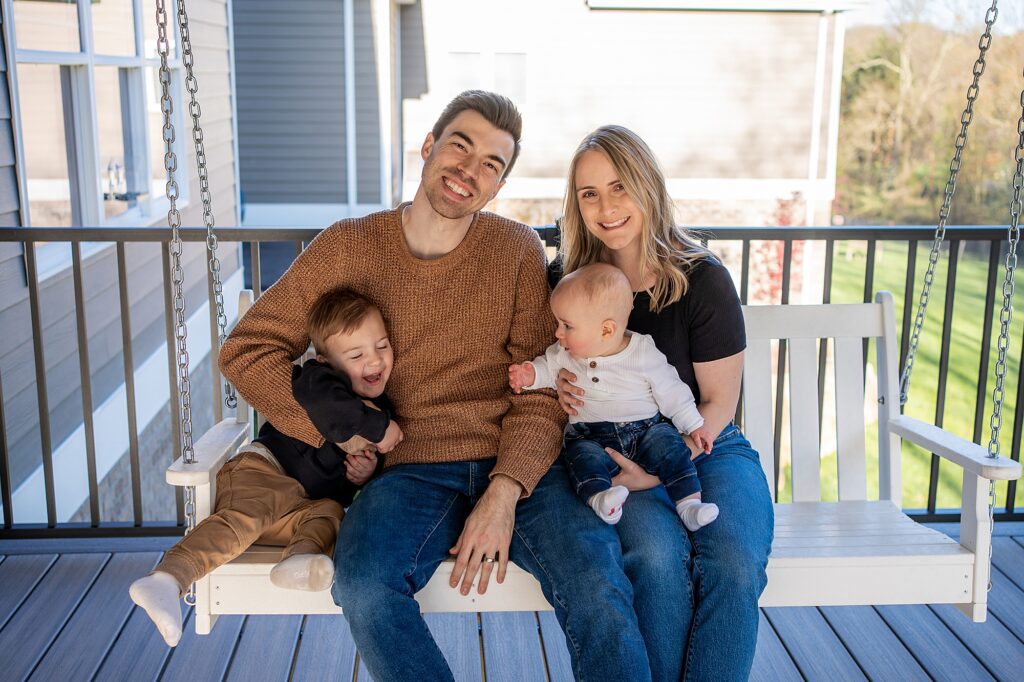 A family of four smiling on a porch swing; the parents, a toddler, and a baby, all enjoy a sunny day.