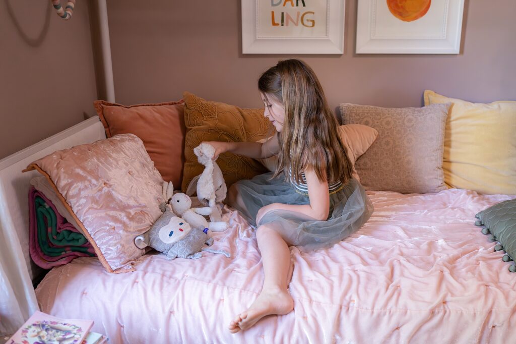 A young girl with long hair, wearing a dress, sits on a bed with pink sheets and colorful pillows, playing with stuffed animals. Two framed pictures hang on the wall above the bed.