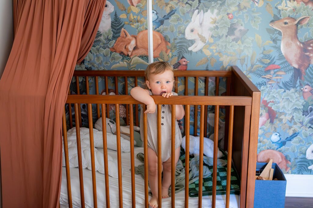 A baby stands in a wooden crib, holding the railing and looking forward. The crib is surrounded by colorful bedding and set against a nature-themed wallpaper with animals and foliage.