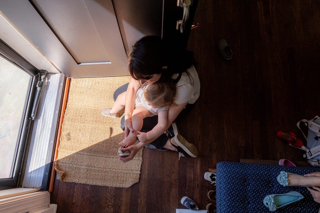 A person sits on the floor by an open door, holding a child on their lap, surrounded by scattered shoes and a woven mat.