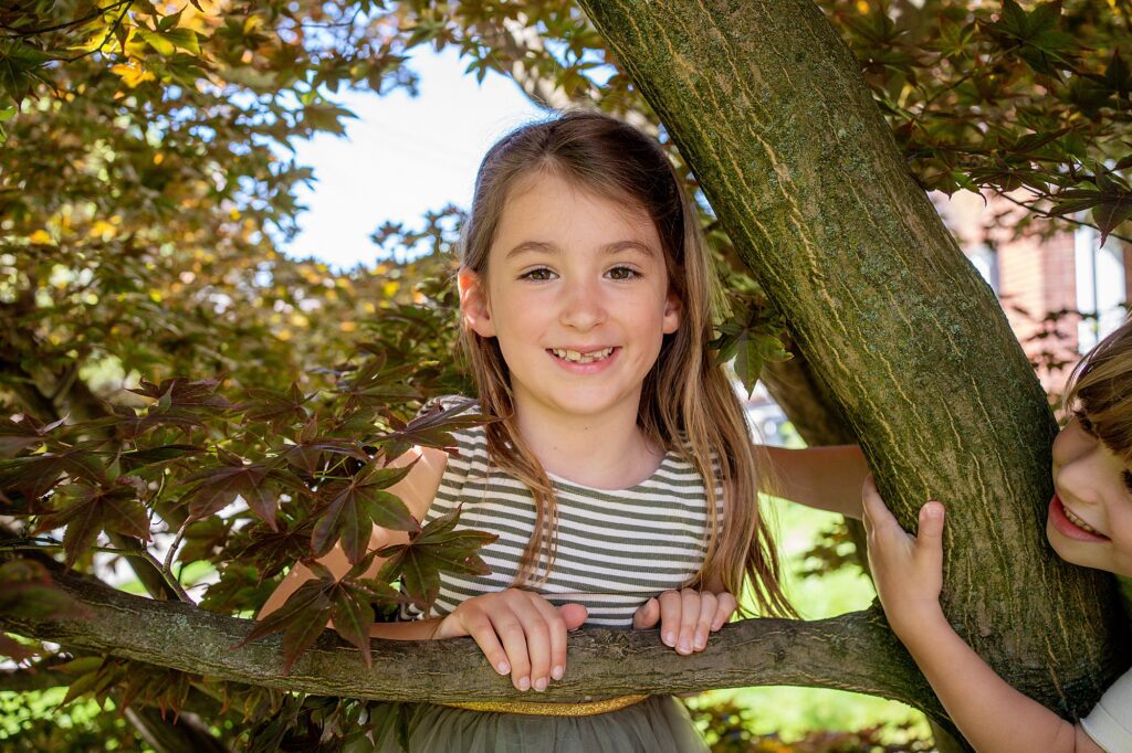 A young girl with long hair and striped shirt stands among tree branches, smiling at the camera. Brown and green leaves surround her. A partially visible child is beside her.
