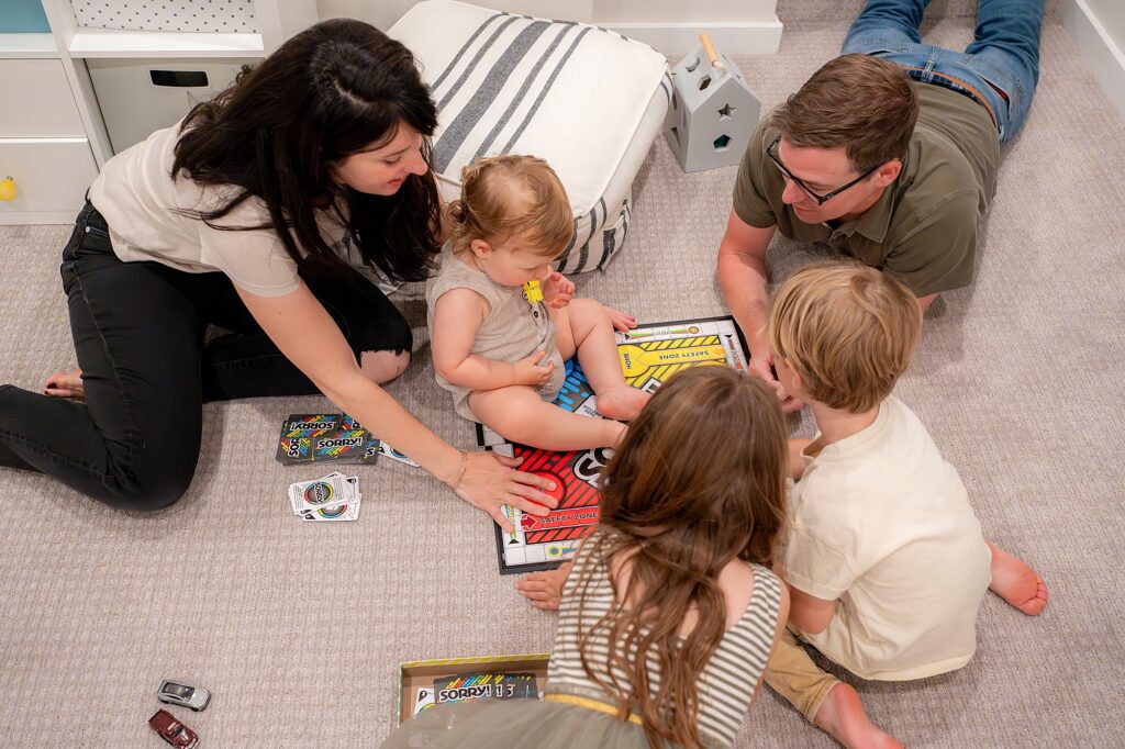 A group of people, including a child and an adult, are playing a board game on the floor in a carpeted room. Toys and game pieces are scattered around them.
