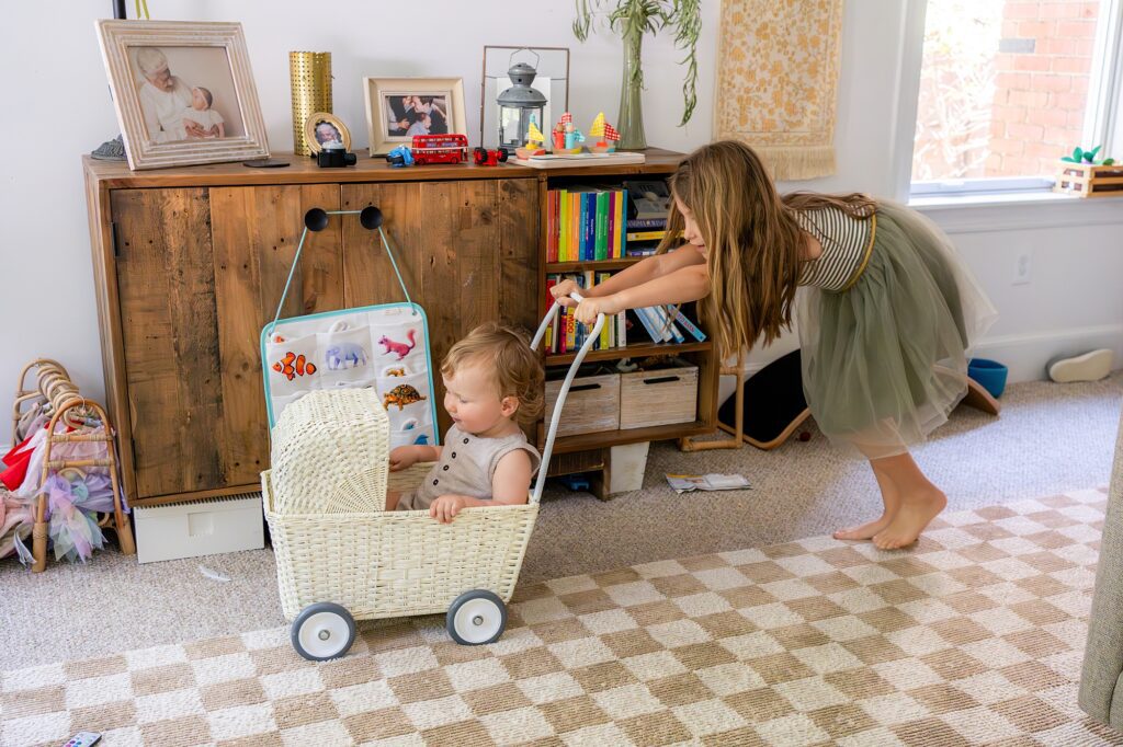 A young girl pushes a baby seated in a wicker cart inside a brightly lit living room. The room has a cabinet with books and toys, and a checkered carpet on the floor.
