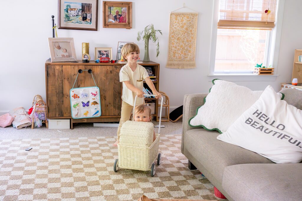 Two children happily play in a living room. One child, pushing a wicker cart, smiles while the other sits inside the cart. The room features a cozy sofa with a "HELLO BEAUTIFUL" pillow and wall decor.