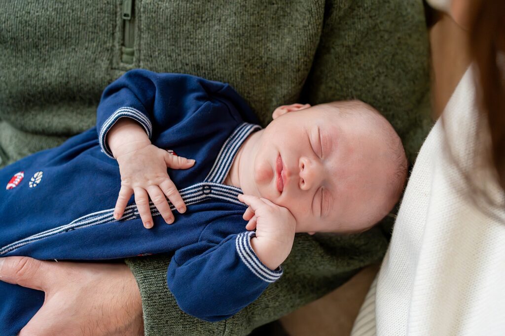 Newborn baby in a blue outfit sleeps peacefully, cradled in an adult's arms wearing a dark green sweater.
