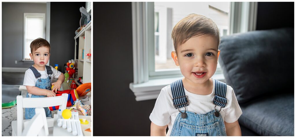 A young child with short brown hair is wearing a white shirt and denim overalls. The child is standing indoors near toys and a gray sofa.