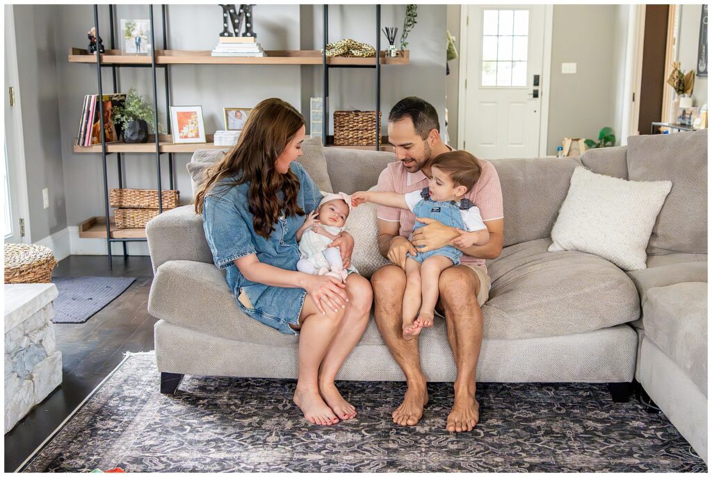 A family of four sits on a sofa. The mother holds a baby, while the father holds a toddler. The toddler reaches towards the baby. A bookshelf and door are in the background.