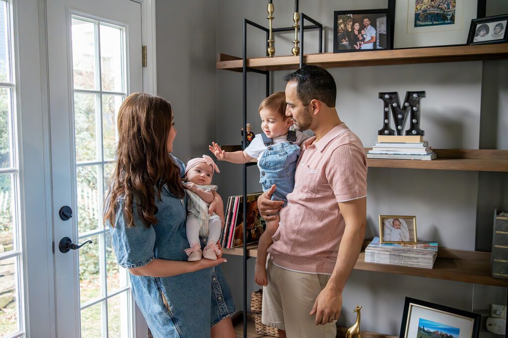 A couple stands by shelves, each holding a child. The woman holds a baby, while the man holds a toddler who reaches towards the baby. Decor and photos fill the shelves beside them.
