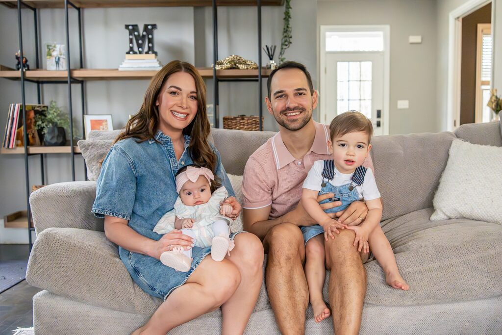 A family of four sits on a couch. The mother holds an infant, and the father holds a toddler. Shelves with decor are in the background.