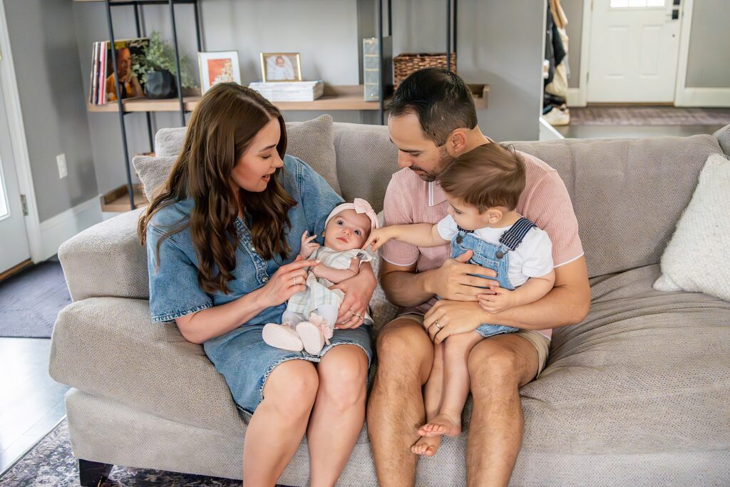 A family of four sits on a couch. The mother holds a baby, and the father holds a young child. The room has photos and decor in the background.