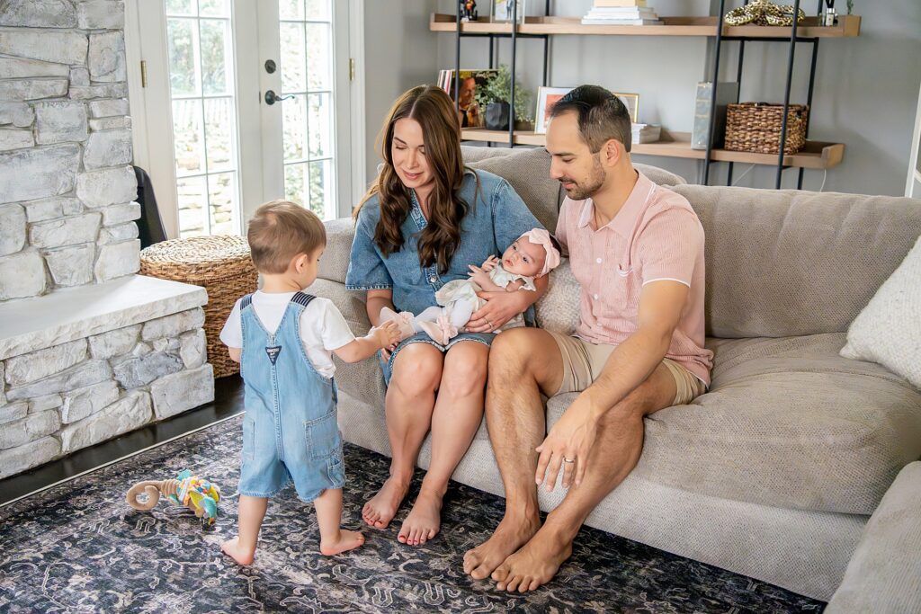 A family sits on a couch in a living room. A woman holding a baby and a man are seated, while a toddler stands nearby. Toys are on the floor, and shelves are in the background.