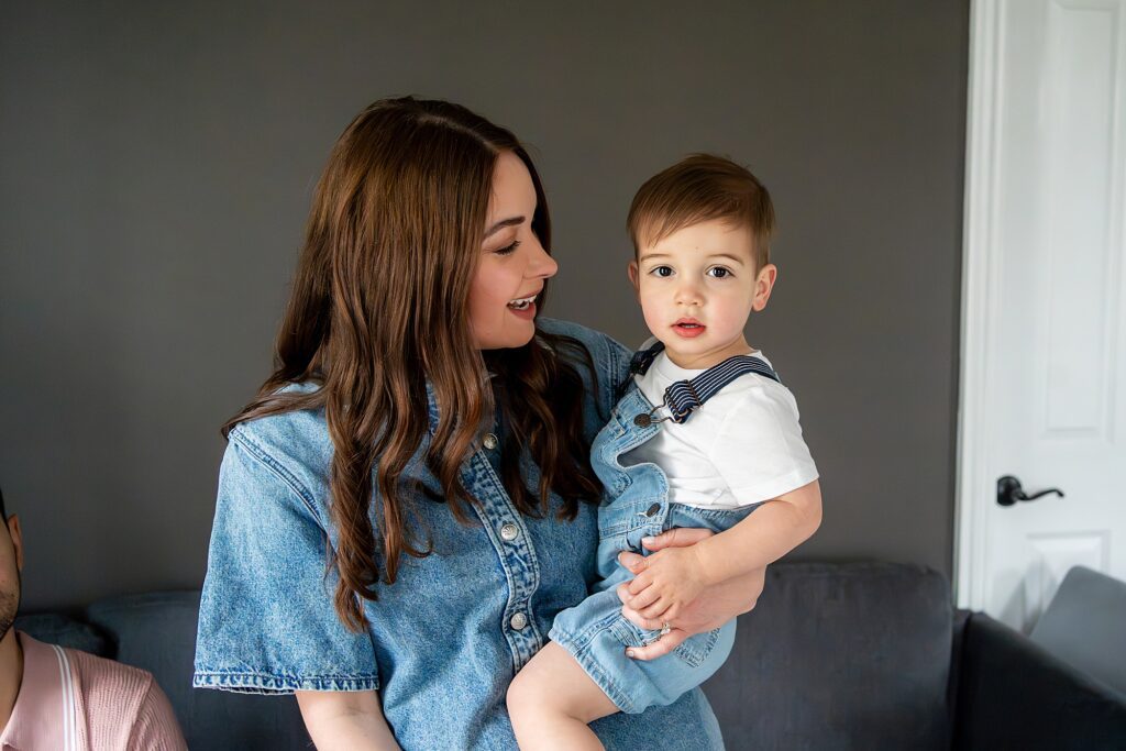 A woman in a denim shirt is holding a young child wearing overalls. They are indoors in front of a gray wall.