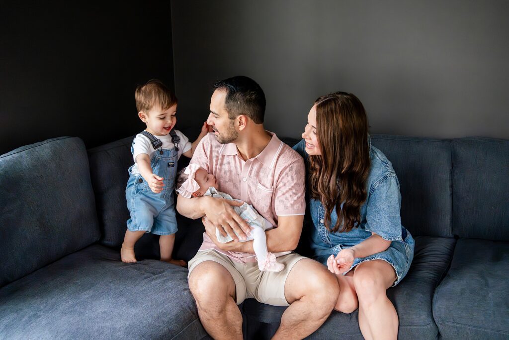 A family sits on a couch. An adult holds a baby while a child stands nearby. Another person sits beside them, smiling.