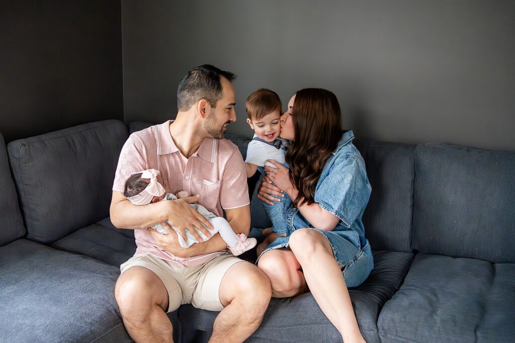 A family sitting on a couch: a man holding a baby, a woman kissing a young child, in casual attire against a gray background.