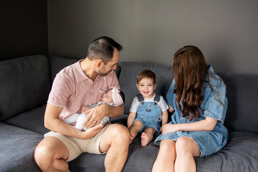 A man and woman sit on a couch with two children; the man holds a baby, and the woman sits with a toddler smiling at the camera.