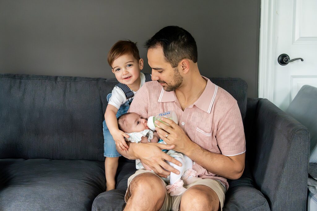 A man sits on a couch feeding a baby from a bottle while a young child leans on his shoulder, smiling at the camera.
