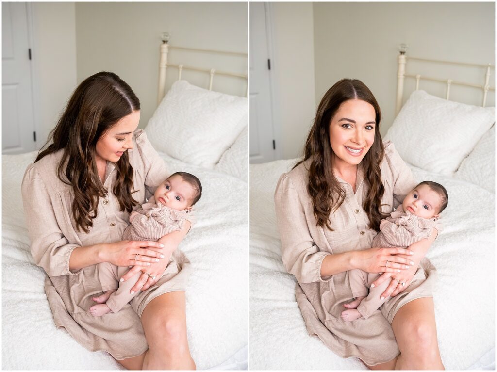 A woman wearing a beige dress is sitting on a bed, holding a baby in her arms. The baby is also dressed in beige. The setting is a neutral-toned bedroom.