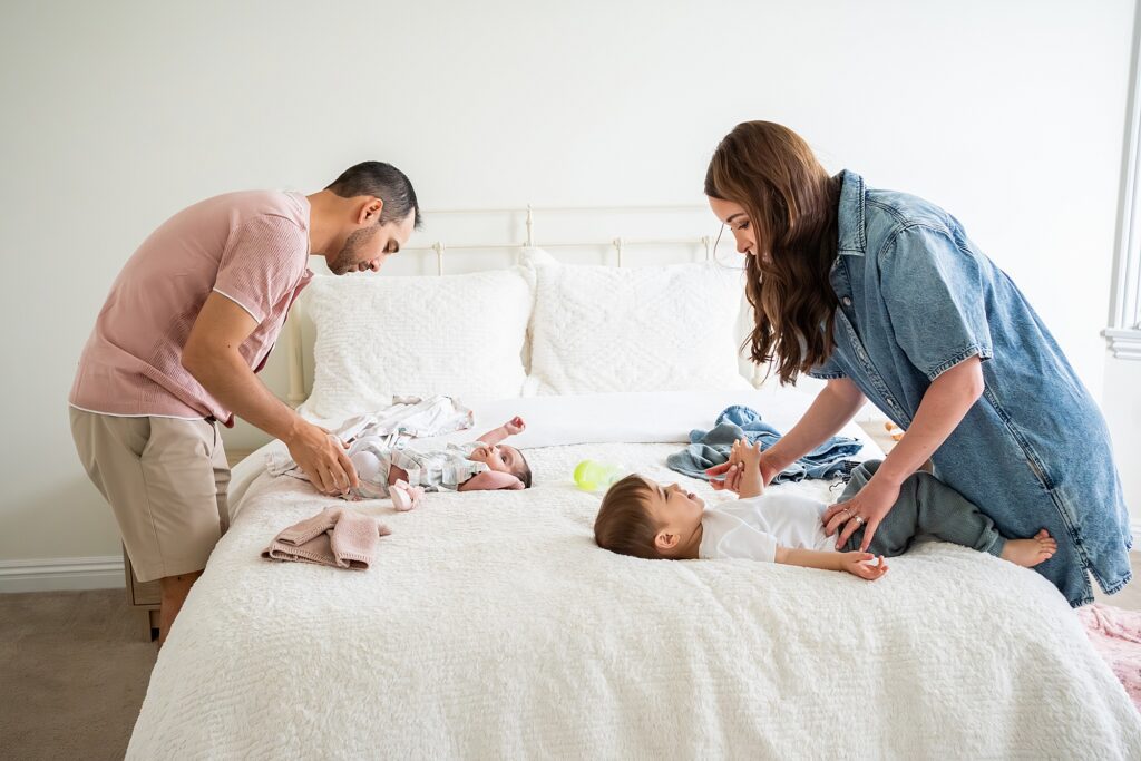 A man and a woman attend to two infants on a bed. The man is changing the baby on the left, and the woman is playing with the baby on the right.
