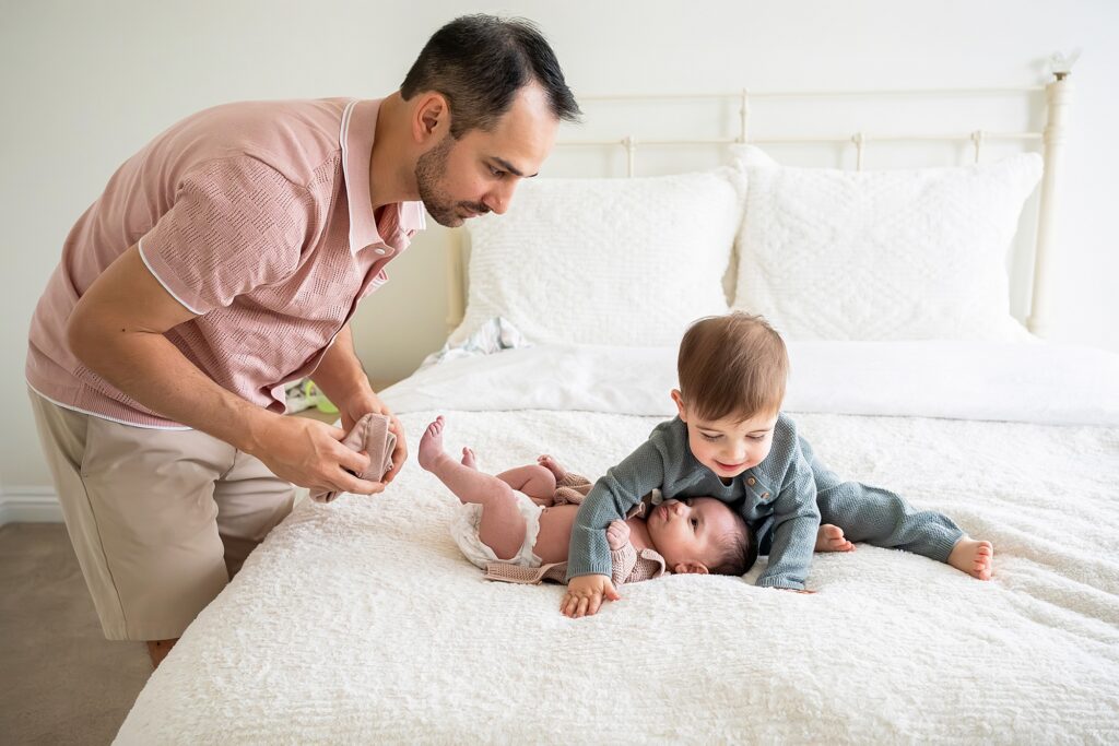 A man prepares to diaper a baby lying on a bed, while a toddler rests their head on the baby's stomach, both wearing comfortable clothing.