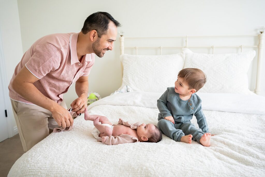 A man plays with a baby lying on the bed while a toddler sits nearby, looking on. The setting is a bedroom with white bedding.