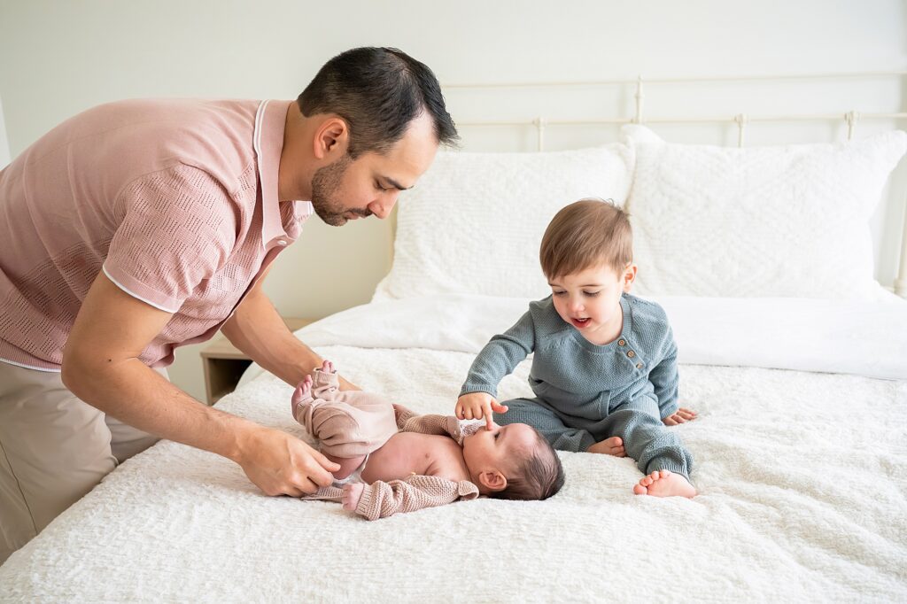 A man tends to a baby in a beige outfit on a white bed. A young child in gray sits nearby, gently touching the baby's face.