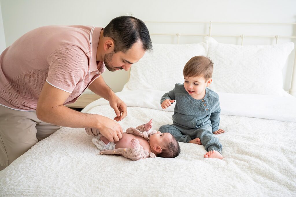 A man adjusts a baby's sweater on a bed, while a toddler in a gray outfit sits beside them, watching. The scene takes place in a bright room with white bedding.