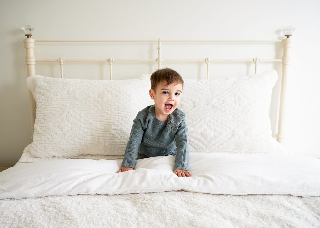 A toddler in a gray outfit is sitting on a white bed with textured pillows, looking forward with an open mouth expression.