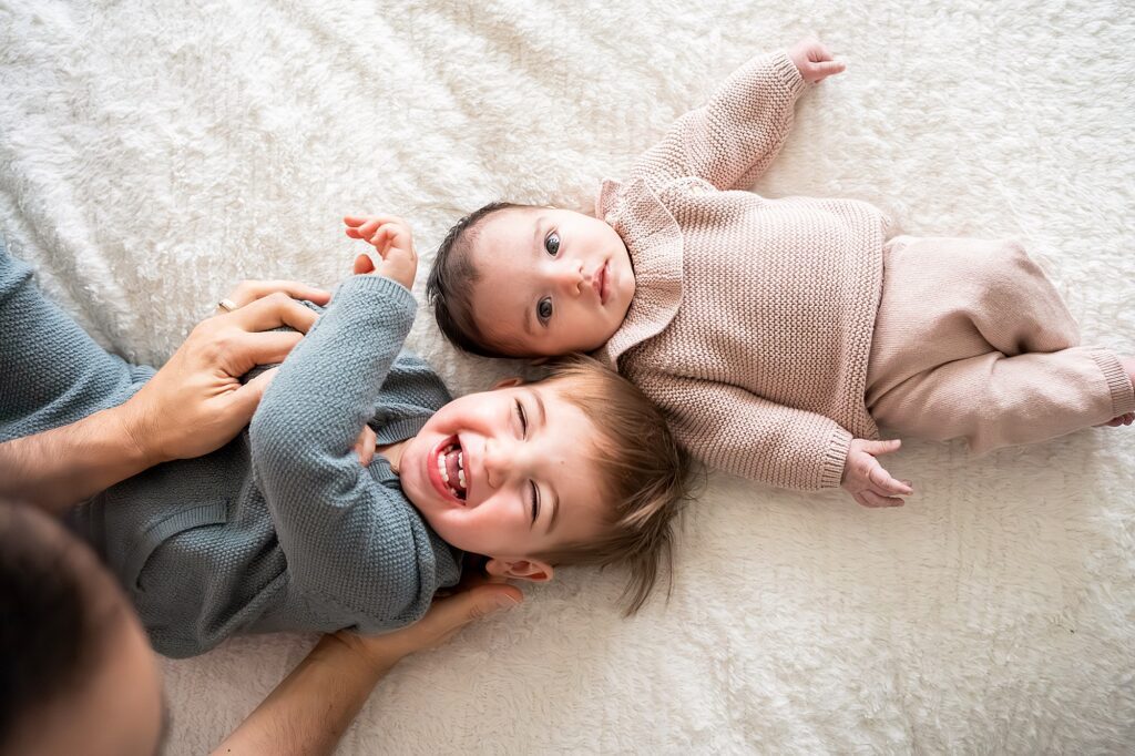 Two children lying on a soft surface. One child is smiling, while the other looks at the camera. An adult's hands are seen tickling the smiling child. Both are dressed in knit outfits.