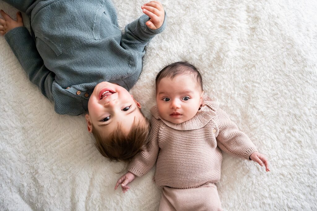 A toddler in a gray sweater lies next to a baby in a pink onesie on a soft white surface, both looking upwards.