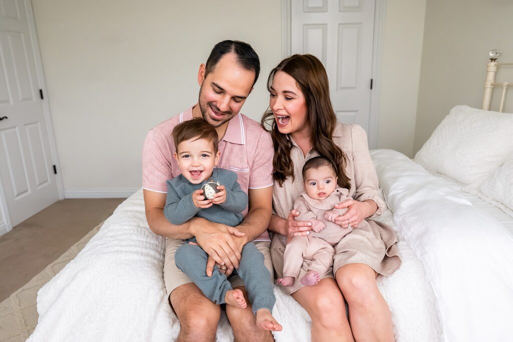 A family of four sits on a bed. The father holds a toddler, and the mother holds an infant. Everyone is smiling. The room is well-lit with white bedding and walls.