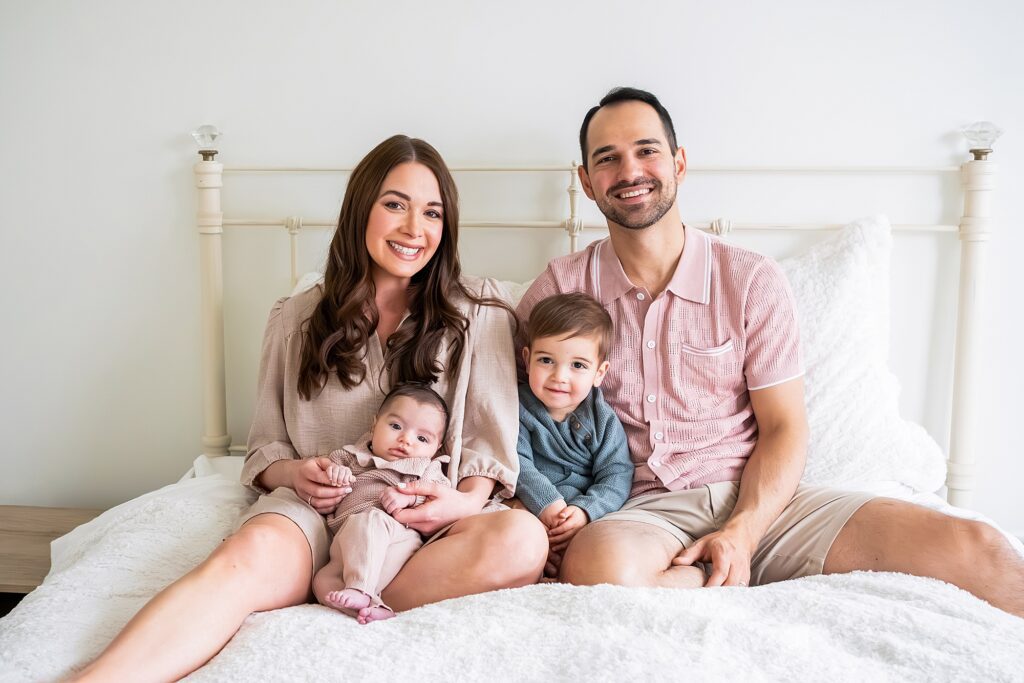 Family sitting on a bed, with a woman holding a baby on her lap and a young child sitting between a man and woman, all smiling at the camera.