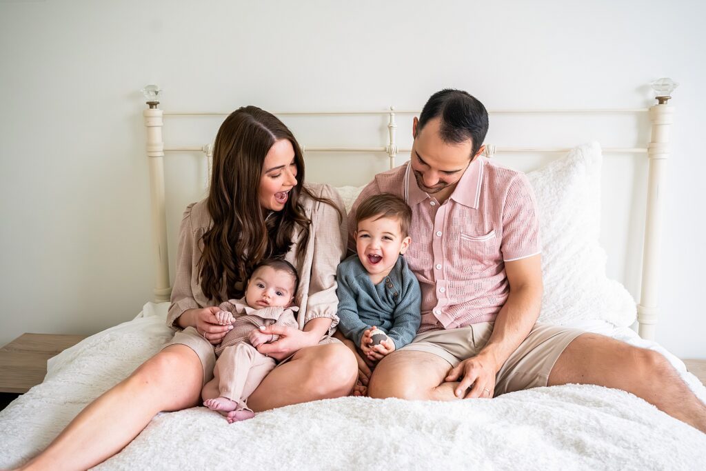 A family sits on a bed: a woman holding a baby, a young child laughing, and a man. They are all smiling and wearing casual, neutral-colored clothing.