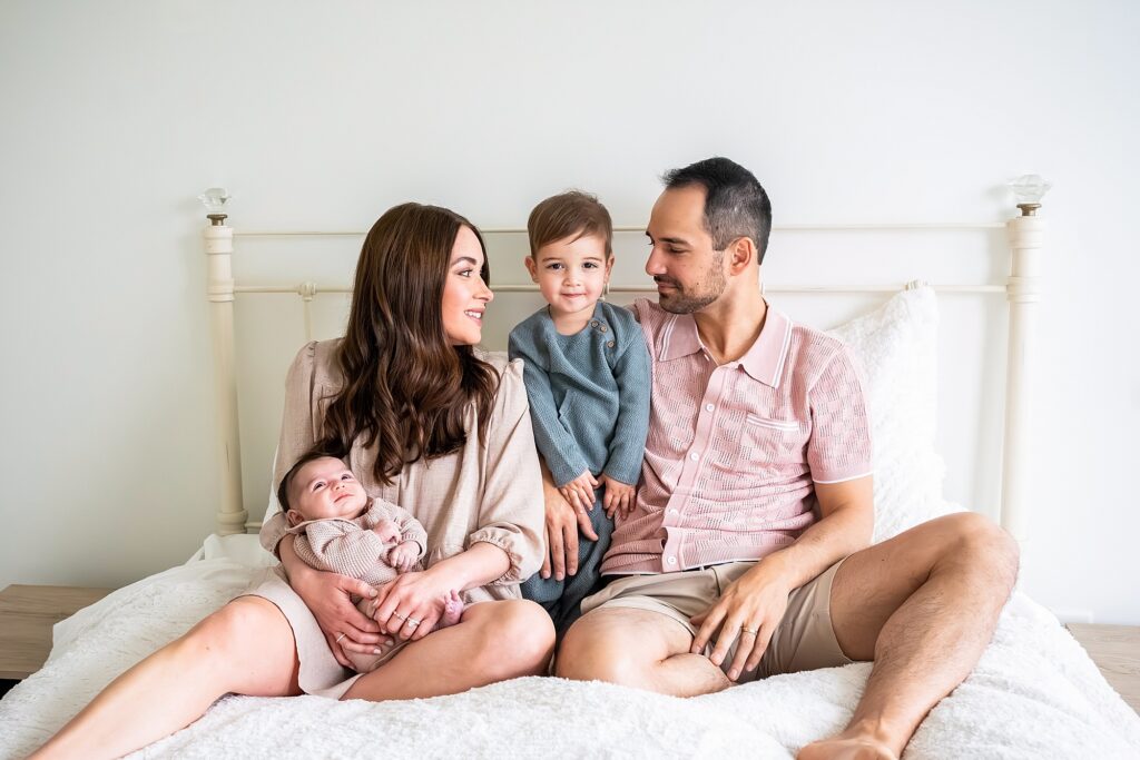 A family of four sits on a bed. A woman holds a baby, a child sits between her and a man, and they are all looking at each other.