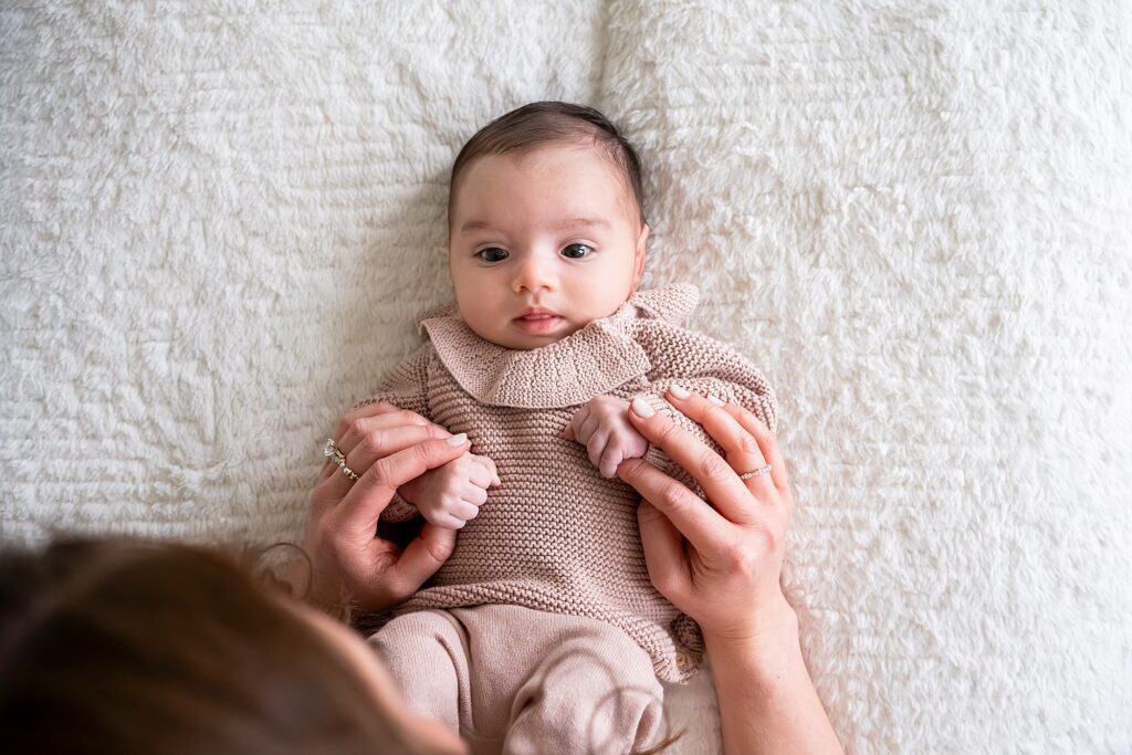A baby lying on a soft surface, wearing a pink knitted outfit. An adult's hands gently hold the baby's hands.