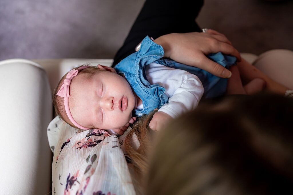 A baby wearing a pink headband and blue outfit sleeps on an adult’s chest, who is seated on a floral-patterned chair.