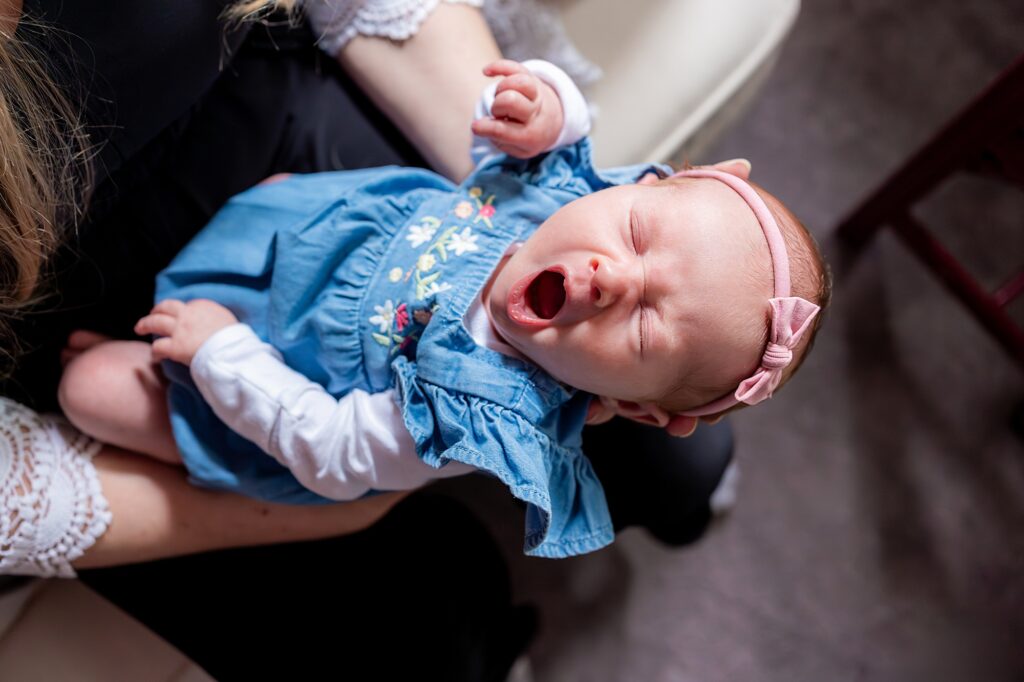 A baby wearing a blue dress and pink headband yawns while being held on an adult's lap.