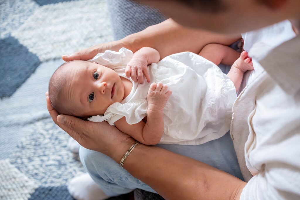 An adult cradles a newborn baby dressed in a white outfit, looking up. The background shows a patterned fabric surface.