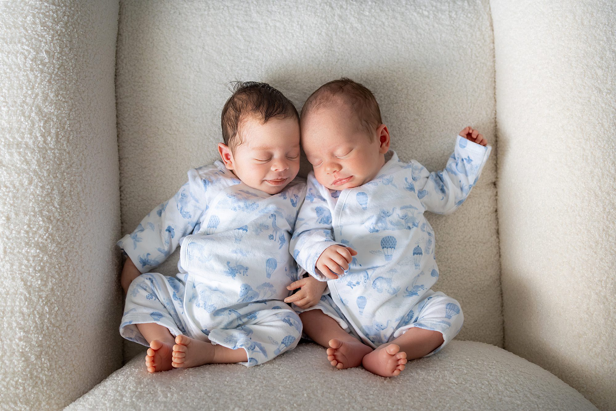 Two newborn babies wearing matching blue and white onesies are sleeping side by side on a soft, light-colored chair.