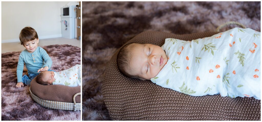 A young boy sits on a brown rug, gently touching the head of a swaddled newborn baby lying on a brown cushion.