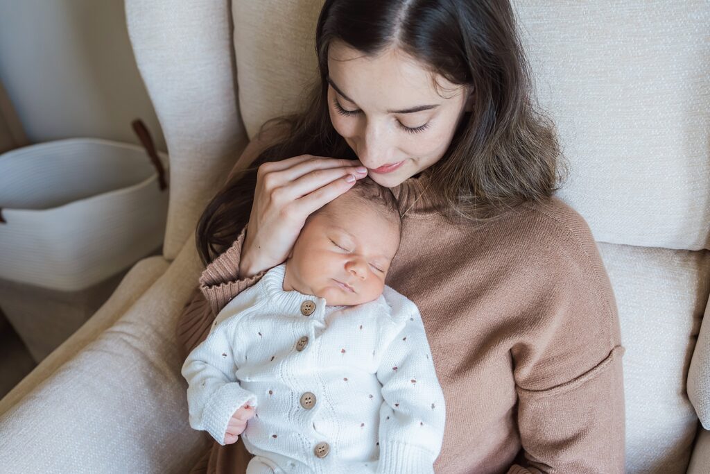 A woman sits in a chair holding a sleeping baby in her arms, gently touching the baby's head.