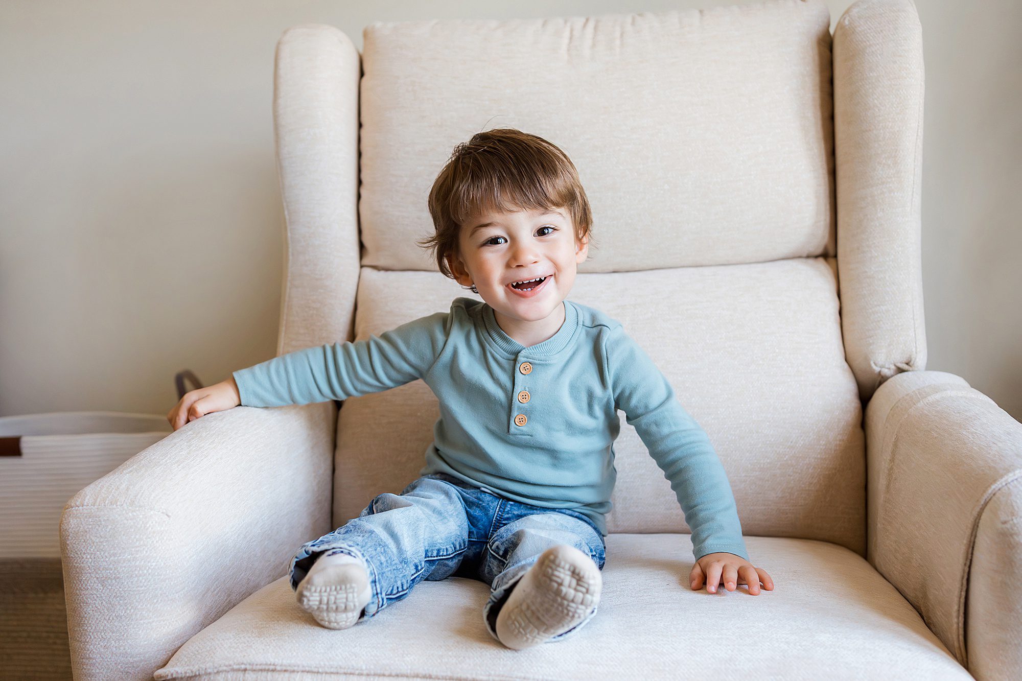 A young child with brown hair, wearing a blue long-sleeve shirt and jeans, sits smiling on a beige armchair.