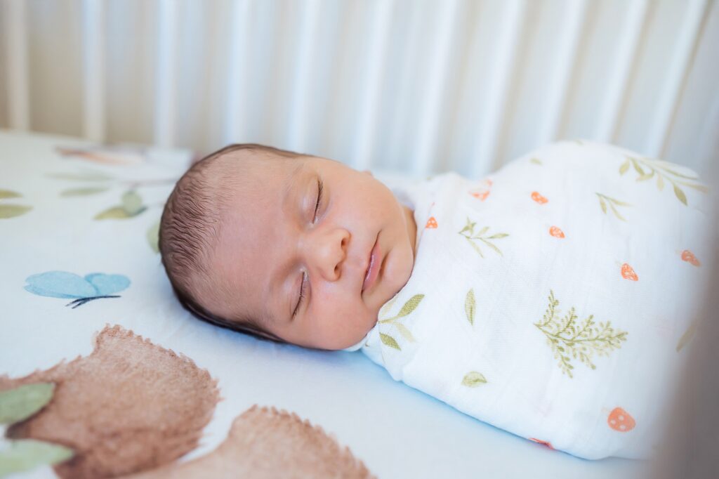 A newborn baby wrapped in a white patterned blanket sleeps peacefully on a crib mattress with animal and plant designs.