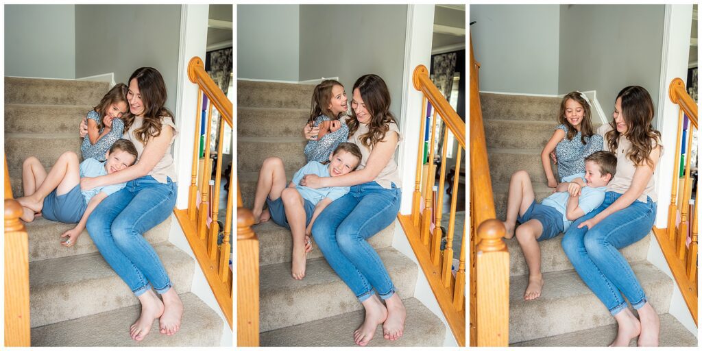 A woman sits on carpeted stairs, smiling and playing with two laughing children, a girl and a boy, in a well-lit home.