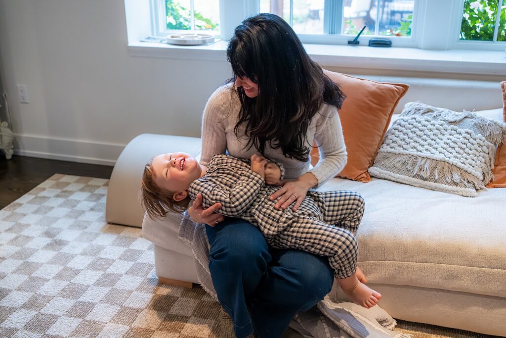 A woman sits on a couch holding a laughing child in plaid pajamas on her lap, both smiling in a living room with pillows and a window in the background.