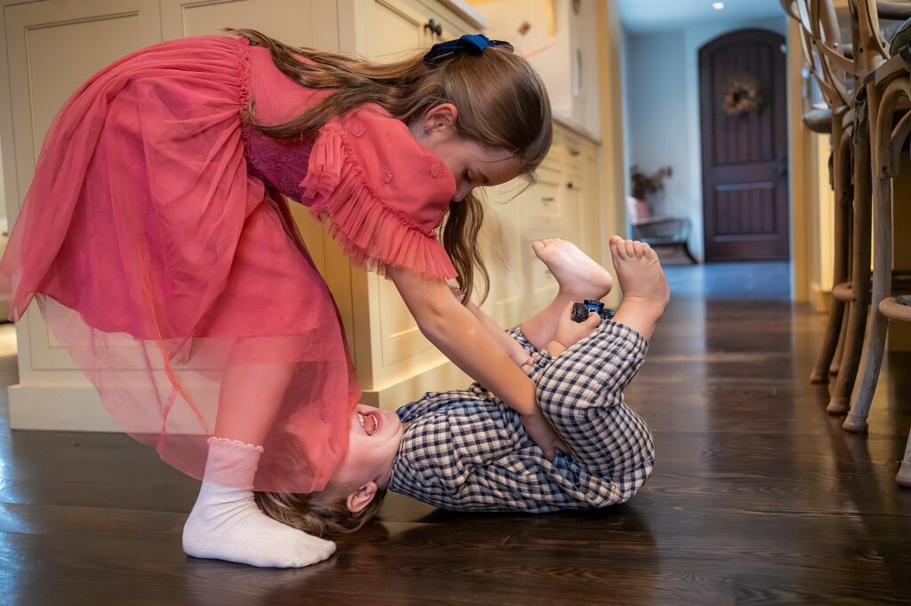 A young girl in a pink dress playfully holds a smiling toddler in checkered pajamas, who is lying on the wooden floor in a kitchen.