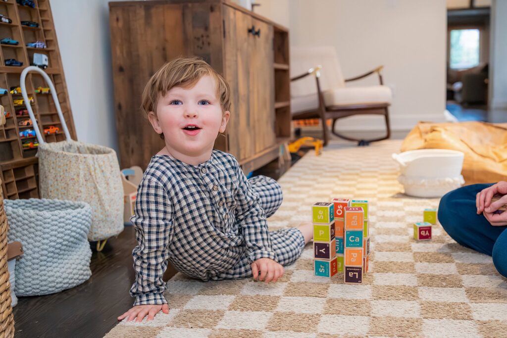 A young child in checkered pajamas sits on a patterned rug, looking up while playing with colorful alphabet blocks indoors.