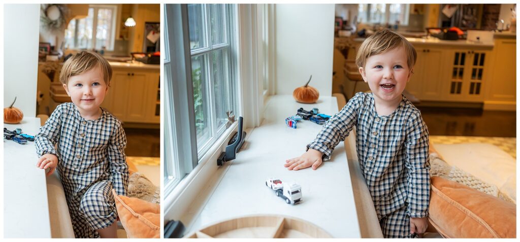 A young child in plaid pajamas stands and smiles while playing with toy cars on a white counter in a kitchen.