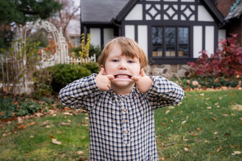 A young child in a plaid shirt stands on a lawn, pulling the sides of their mouth to make a wide, exaggerated smile. A house with a Tudor-style facade is in the background.